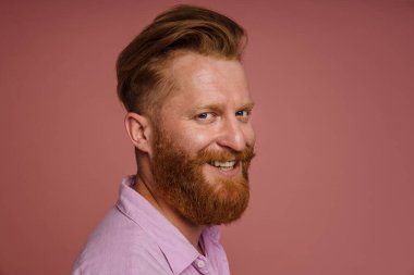 Portrait of adult handsome stylish redhead bearded smiling man in pink shirt looking at camera , while standing over isolated pink background