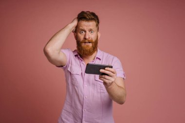 Adult stylish redhead bearded confused man with phone holding his head and looking at camera , while standing over isolated coral background