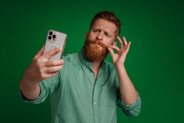 Ginger white man gesturing while taking selfie on mobile phone isolated over green background
