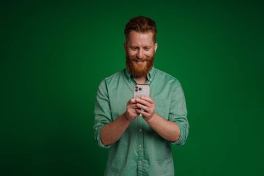 Ginger white man with beard smiling and using mobile phone isolated over green background