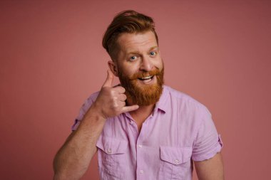 Ginger white man with beard smiling and making handset gesture isolated over pink background