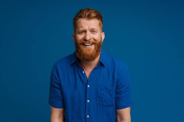 Ginger white man with beard smiling and looking at camera isolated over blue background
