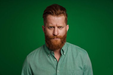Ginger white man with beard frowning and looking at camera isolated over green background