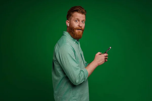 Ginger Shocked Man Earphones Using Mobile Phone Isolated Green Background — Stock Photo, Image