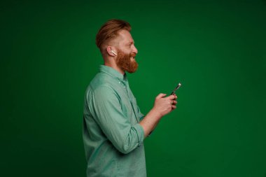 Ginger white man in earphones smiling and using mobile phone isolated over green background