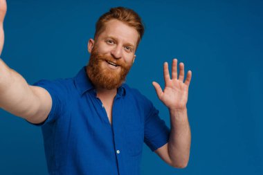 Ginger white man with beard gesturing while taking selfie photo isolated over blue background