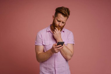 Ginger white man with beard frowning and using mobile phone isolated over pink background