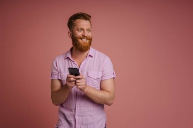 Ginger white man with beard smiling and using mobile phone isolated over pink background