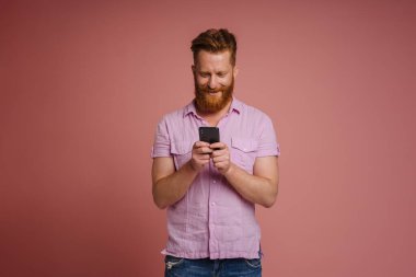Ginger white man with beard smiling and using mobile phone isolated over pink background