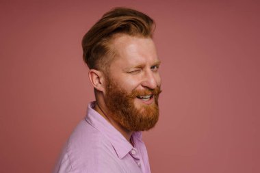 Ginger white man with beard winking and smiling at camera isolated over pink background