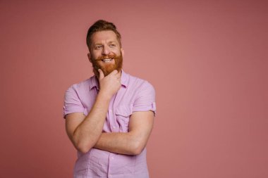 Ginger white man with beard smiling and holding his chin isolated over pink background