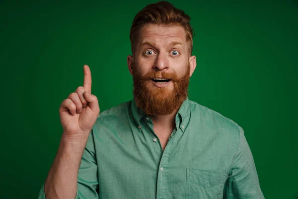 Ginger white man expressing surprise and ointimg finger upward isolated over green background