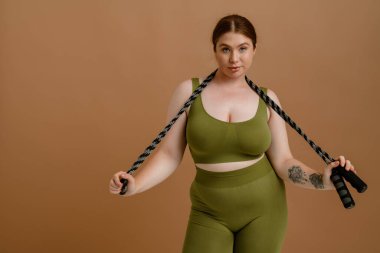 Young beautiful calm plus size girl holding jumping rope on her neck both hands and looking at camera , while standing over isolated brown background clipart