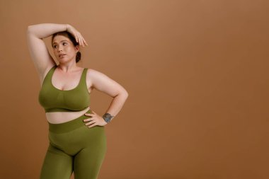 Güzel, sakin, uzun boylu, antrenman elbiseli bir kız. Kolları başının üzerinde, tek başına kahverengi arka planda dikilirken yan bakıyor.