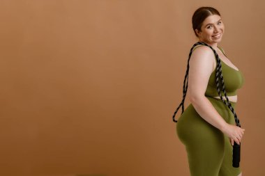 Young beautiful smiling happy plus size girl holding jumping and looking at camera , while standing with her side over isolated brown background clipart