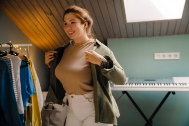 Genç, güzel, gülümseyen, mutlu bir kız gömleğini giyip yan tarafa bakıyor. Evdeki rahat odada dikiliyor.