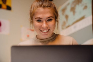 Evde otururken dizüstü bilgisayara bakıp gülümseyen güzel bir kızın portresi.