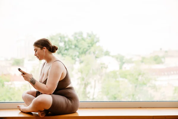 stock image Ginger young woman listening music and using cellphone after yoga practice indoors