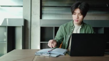 Kendine güveni tam Asyalı bir adam laptopta çalışıyor ve çalışma odasındaki masada yazı yazıyor.