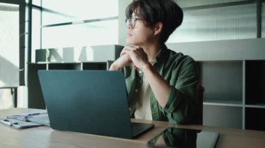 Ciddi esmer, gözlüklü Asyalı bir adam çalışma odasındaki bir masada dizüstü bilgisayarla çalışıyor.