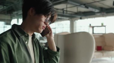 Çalışma odasında gözlüklü üzgün bir Asyalı telefonda konuşuyor.