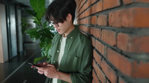 一位戴眼镜的友善的亚洲男人在一间联合工作间的砖墙边用智能手机发短信 — 图库视频影像