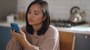 Pozitif Asyalı kadın biriyle konuşuyor ve evdeki mutfakta cep telefonuyla yazıyor.