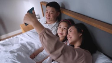Neşeli Asyalı aile evde yatakta yatarken cep telefonunda selfie çekiyorlar.