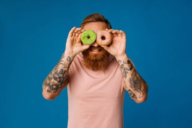 Sakallı mutlu adam mavi arka planda tek başına dururken donut tutuyor.