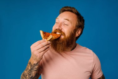Sakallı mutlu adam, mavi arka planda tek başına dikilirken pizza yiyor.