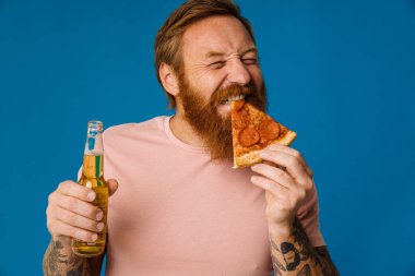 Sakallı mutlu adam elinde pizza dilimi ve bira tutarken mavi arka planda tek başına duruyor.