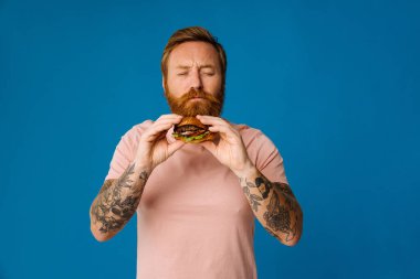 Sakallı, aç bir adam hamburger yerken mavi arka planda tek başına duruyor.