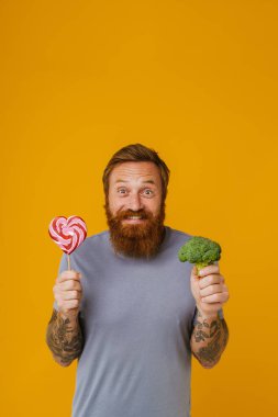 Sakallı adam elinde lolipop ve brokoli tutarken sarı arka planda tek başına duruyor.