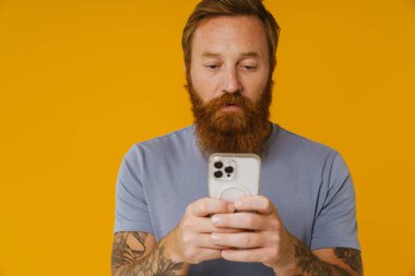 Sakallı beyaz adam, sarı arka planda tek başına dururken elinde cep telefonu tutuyor.