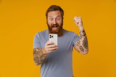 Sakallı beyaz adam, sarı arka planda tek başına dururken elinde cep telefonu tutuyor.