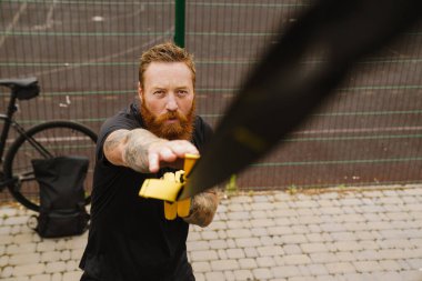 Genç, yakışıklı, sakallı, ciddi, kızıl saçlı bir adam. Açık hava spor sahasında TRX ile egzersiz yapıyor.