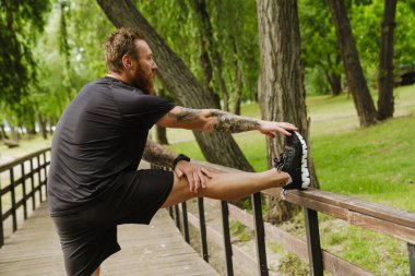 Genç, yakışıklı, gülümseyen, sakallı, sakallı bir adam. Bacağını uzatıyor ve güneşli parktaki ahşap korkuluklara tırmanıyor.