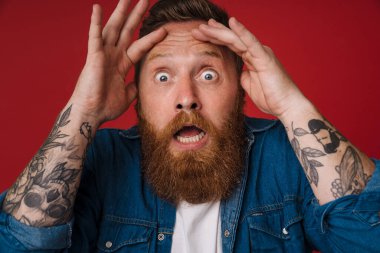 Yetişkin sakallı, yakışıklı, şok olmuş, ağzı açık, elleri alnında kırmızı arka planda dikilirken kameraya bakan kızıl saçlı adam.