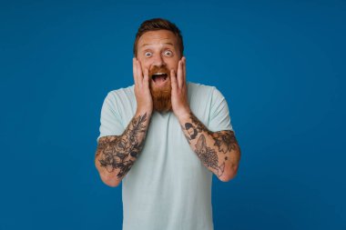 Yetişkin sakallı, yakışıklı, şaşkın, ağzı açık, yanaklarını tutarak kameraya bakan, izole edilmiş mavi arka planda duran adam.