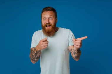 Yetişkin sakallı, yakışıklı, stil düşkünü, ağzı açık, iki eliyle kameraya bakan ve izole edilmiş mavi arka planda duran adam.