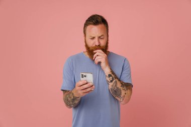 Yetişkin sakallı, yakışıklı, düşünceli bir adamın portresi. Telefonuna bakıyor ve sakalını tutuyor. İzole edilmiş pembe arka planda duruyor.