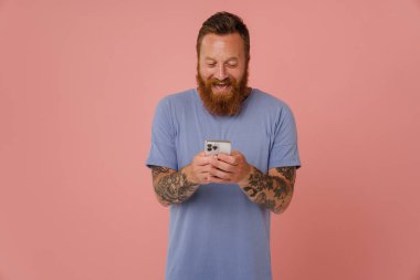 Yetişkin sakallı, yakışıklı, hevesli bir adamın portresi. Telefonunu tutuyor ve bakıyor. İzole edilmiş pembe arka planda duruyor.
