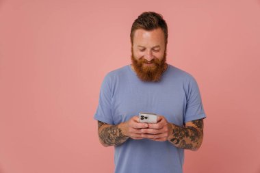 Yetişkin sakallı, yakışıklı, stil sahibi, gülümseyen ve telefonunu kullanan bir adamın portresi.