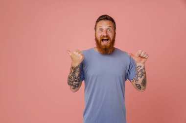 Yetişkin, kızıl saçlı, sakallı, ağzı açık, ağzı açık, baş parmaklarıyla işaret eden, elleri kaldıran ve kameraya bakan, pembe arka planda duran hevesli bir adam.