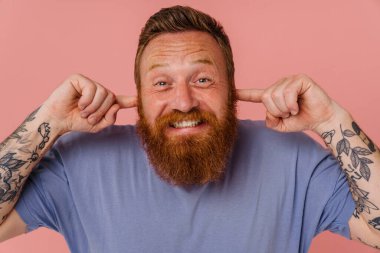 Yetişkin kızıl saçlı, sakallı, tişörtlü, dişlerini sıkmış, kulaklarını parmaklarıyla tıkayan pembe arka planda dikilip kameraya bakan gergin adam.
