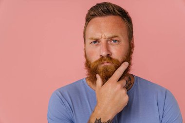 Kızıl saçlı, sakallı bir adam pembe arka planda yalnız başına poz veriyor ve kameraya bakıyor.