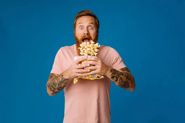 Shocked bearded white man in casual wear holding bucket with flying popcorn isolated over blue studio background