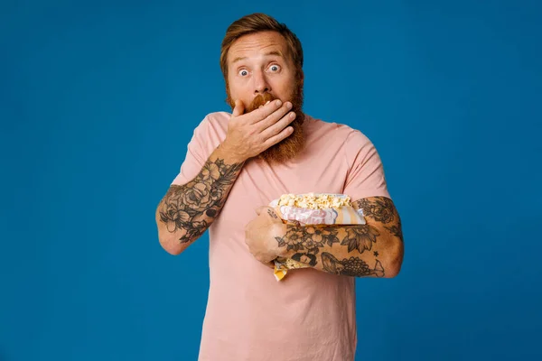 Shocked bearded white man in casual wear eating popcorn isolated over blue studio background