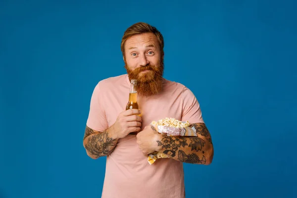 Pleased ginger bearded man holding beer and popcorn while standing isolated over blue studio background