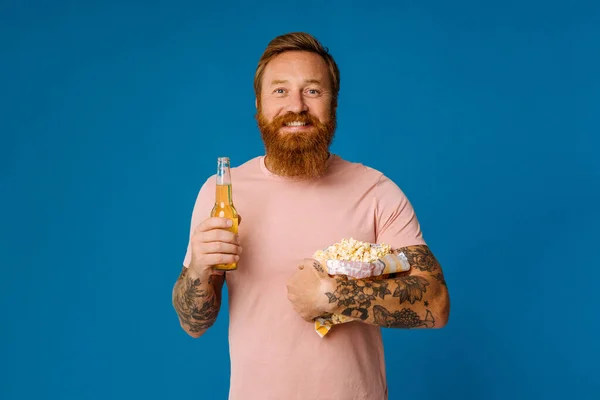 Portrait of excited ginger bearded man holding beer and popcorn while standing isolated over blue studio background